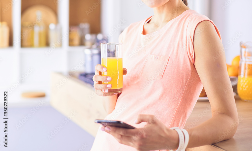 Smiling pretty woman looking at mobile phone and holding glass of orange juice while having breakfast in a kitchen. Smiling pretty woman looking at mobile phone and holding glass of orange juice while having breakfast in a kitchen.