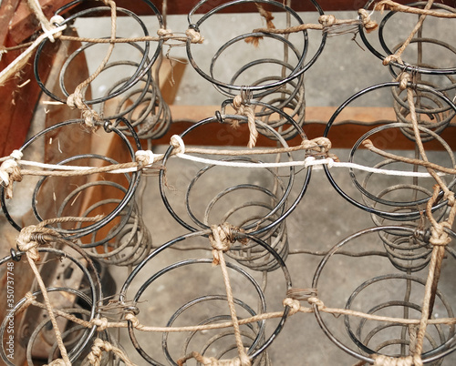 Rusted metal wire chair coils exposed in an antique wooden chair seat. Rows of metal spirals with jute twine rope. Laid out in an array of pattern. Chair is a vintage rocker.