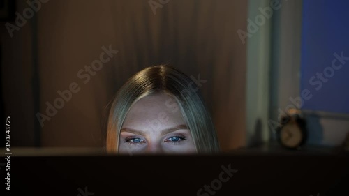 Crop view of beautiful female sitting at night at computer and looking at camera over top of monitor with dark window on blurred background