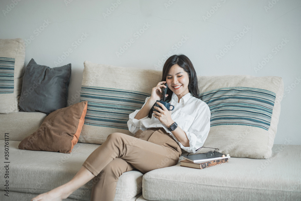 Fototapeta premium Pretty young asian women talking on the phone and holding coffee on the sofa at home.