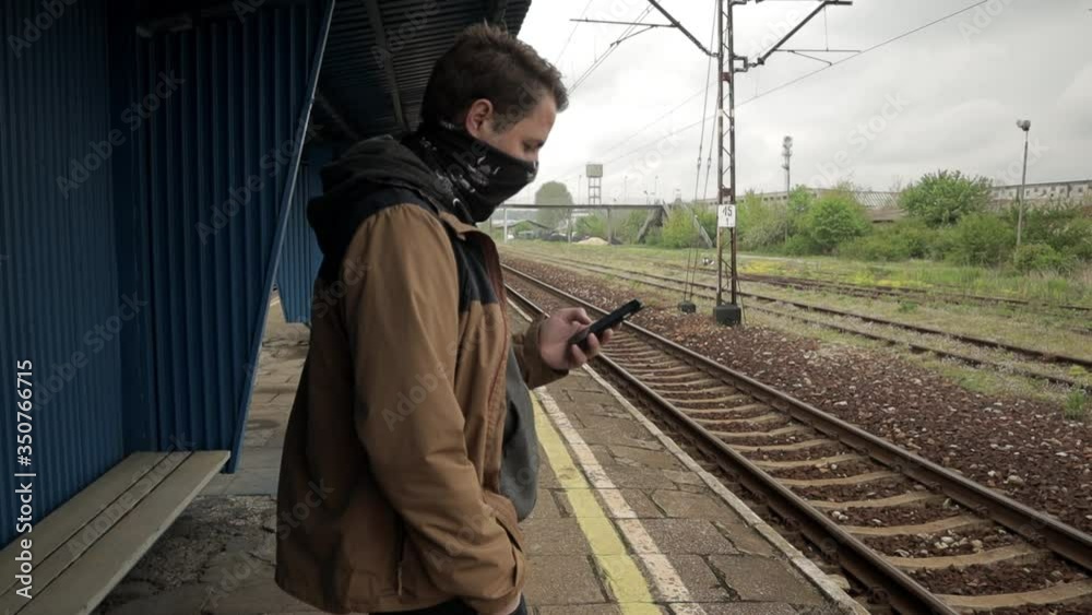 Young man with face covered is checking his mobile phone on train station platform while waiting on train arrival. One person. 24fps