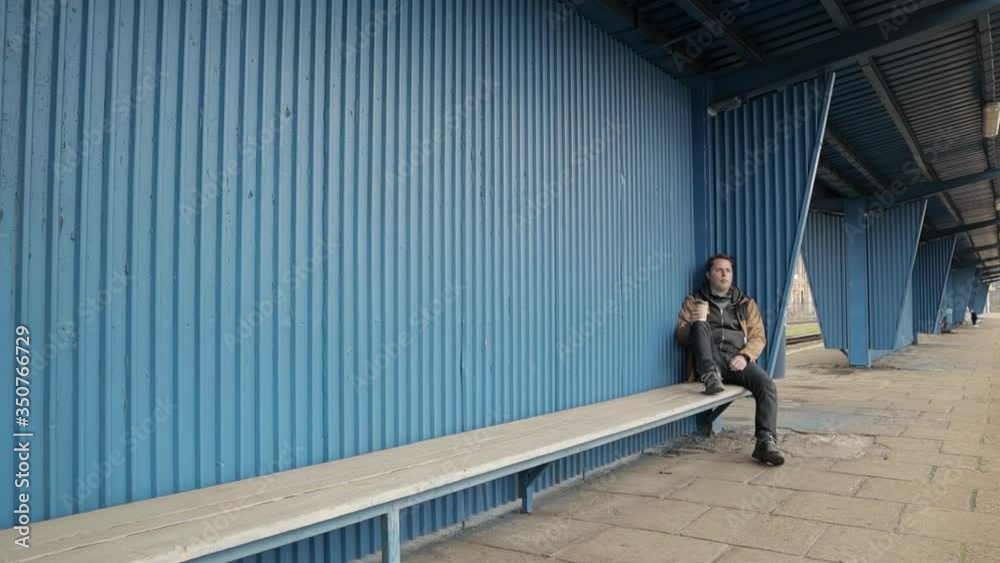 Bored young man sits with one foot on wooden bench, leaning against blue shelter on train station platform and drinks coffee while waiting on train arrival. One person. wide shot, 24fps Bored young man sits with one foot on wooden bench, leaning against blue shelter on train station platform and drinks coffee while waiting on train arrival. One person. wide shot, 24fps