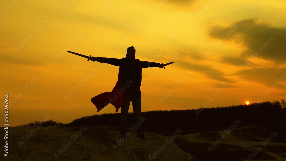a warrior with sword in his hand and in red cloak stands on mountain in sunset light. free male knight prays with swords outstretched hands. free man plays superhero. game of Roman legion.