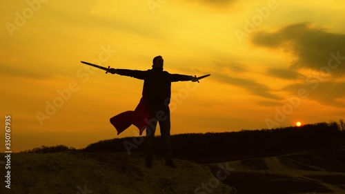 a warrior with sword in his hand and in red cloak stands on mountain in sunset light. free male knight prays with swords outstretched hands. free man plays superhero. game of Roman legion. © zoteva87