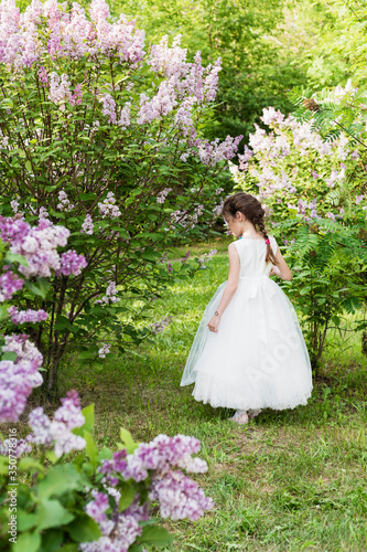 Happy girl in white dress in the park with blooming lilacs, enjoys spring and blooming. Beautiful spring garden, childhood