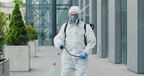 Caucasian man in special antibacterial white suit and respirator walking the street and disinfecting sidewalk with pulverizator. Spraying disinfectant on ground outdoors. Coronavirus pandemic concept. photo