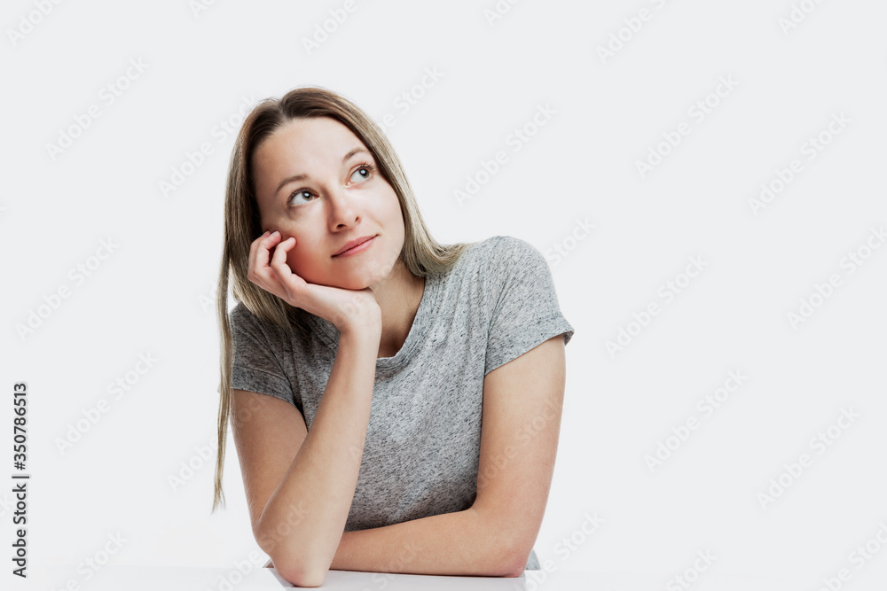 A smiling young woman is sitting and dreamily looking up propping her head with her hand. Dreams and plans. Isolated on a white background. Space for text. A smiling young woman is sitting and dreamily looking up propping her head with her hand. Dreams and plans. Isolated on a white background. Space for text.