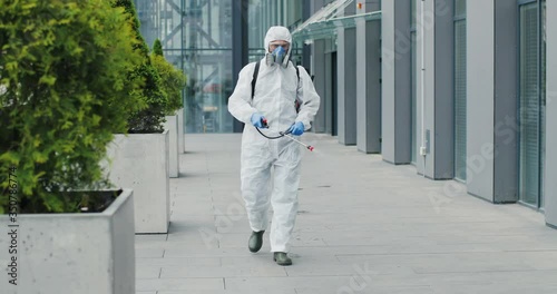 Man in special antibacterial white suit and respirator going by street and disinfecting pavement with pulverizator. Spraying disinfectant on sidewalk outdoors. Covid-19 pandemic outbreak concept. photo