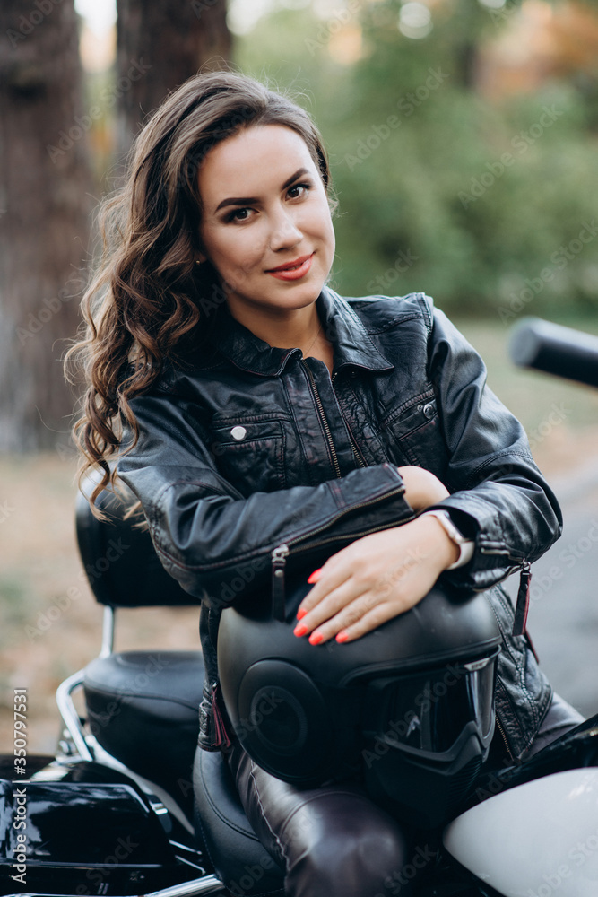 Young beautiful biker woman in a black leather jacket on a motorcycle with a helmet in his hands. The concept of speed and freedom. Soft selective focus.