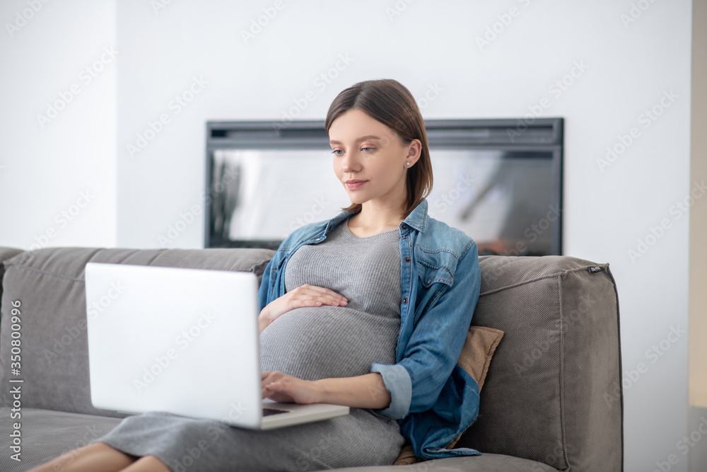 Naklejka premium Young pregnant woman in a jeans jacket watching something online