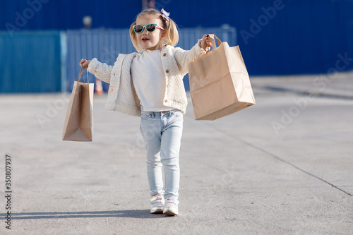 Cheerful baby with purchases. Little girl holding shopping bags.Little girl alone in spring outdoors with paper bags for shopping.Girl in sun glasses posing alone on the street with shopping bags