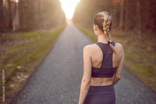 Sporty healthy young woman walking at sunrise along a rural road through a dense forest towards the glow of the sun at the end between the trees in a close up rear view