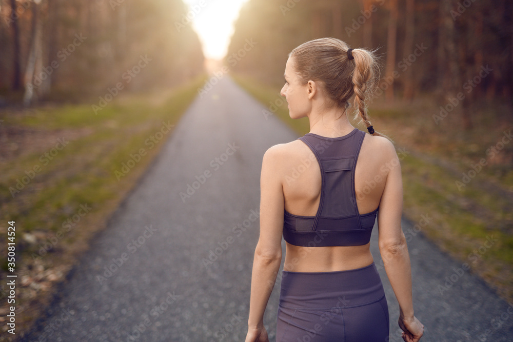 Sporty healthy young woman walking at sunrise along a rural road through a dense forest towards the glow of the sun at the end between the trees in a close up rear view Sporty healthy young woman walking at sunrise along a rural road through a dense forest towards the glow of the sun at the end between the trees in a close up rear view
