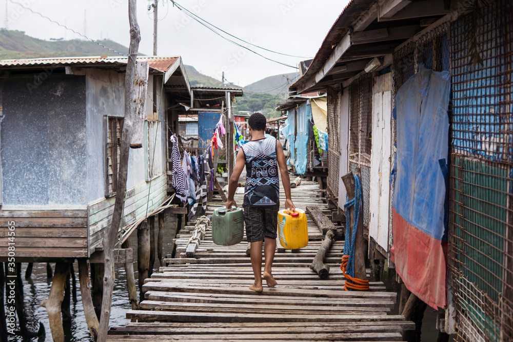 unrecognizable young man carrying jerrycans filled with water walking over wooden path over the water in floating village