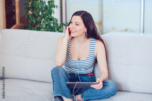 a beautiful woman of mixed race Millennials sits on the sofa at home in a cozy atmosphere with a tablet, smiles, communicates with friends online, receives remote online education at home. 