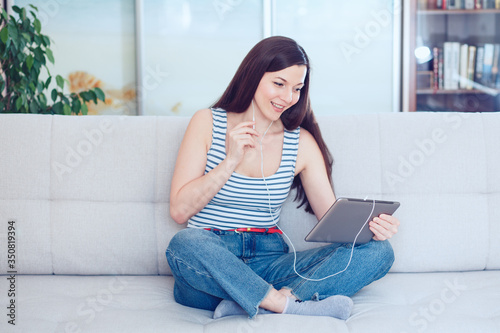 a beautiful woman of mixed race Millennials sits on the sofa at home in a cozy atmosphere with a tablet, smiles, communicates with friends online, receives remote online education at home. 