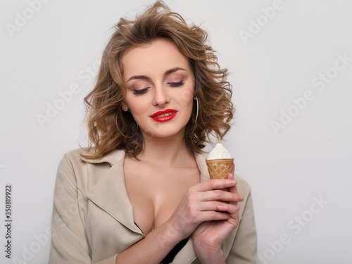 Sexy woman with red lips curly hair in short coat and black bra playfully posing with delicious ice cream on white background with closed eyes