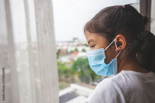 Asian child girl wearing protection mask looking outside through the window and stay at home quarantine from the coronavirus Covid-19 and air pollution pm2.5.