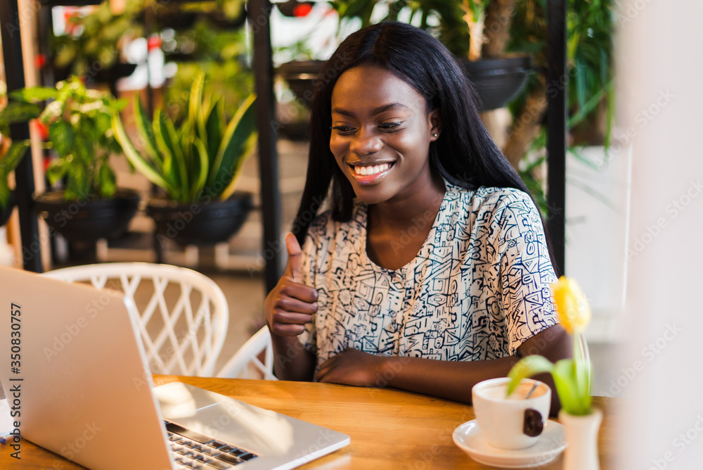 Portrait of an african american young woman with thumbs up sitting at cafe with laptop writing her blog Portrait of an african american young woman with thumbs up sitting at cafe with laptop writing her blog