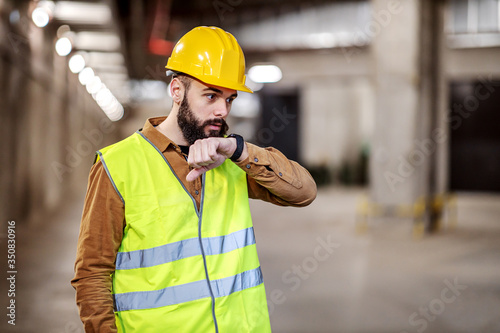 Dedicated bearded supervisor standing in underground garage in construction process and talking with investors over smart watch.