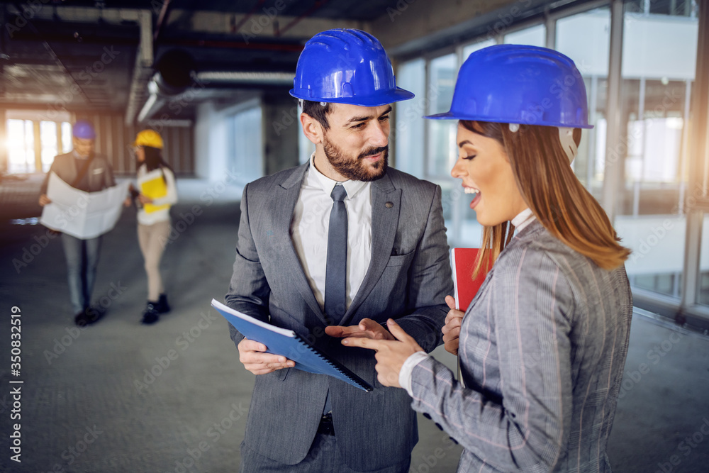 Two smiling attractive architects with helmets on heads standing in building in construction process, holding paperwork and talking about fresh ideas for building renew. Construction site Two smiling attractive architects with helmets on heads standing in building in construction process, holding paperwork and talking about fresh ideas for building renew. Construction site