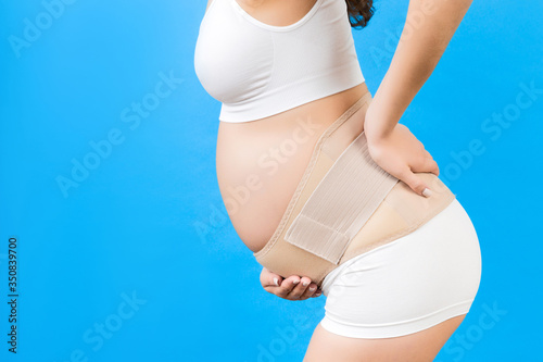 Close up of pregnant woman in underwear with bandage against backache at blue background with copy space. Mother is suffering from pain in the back. Orthopedic abdominal support belt concept