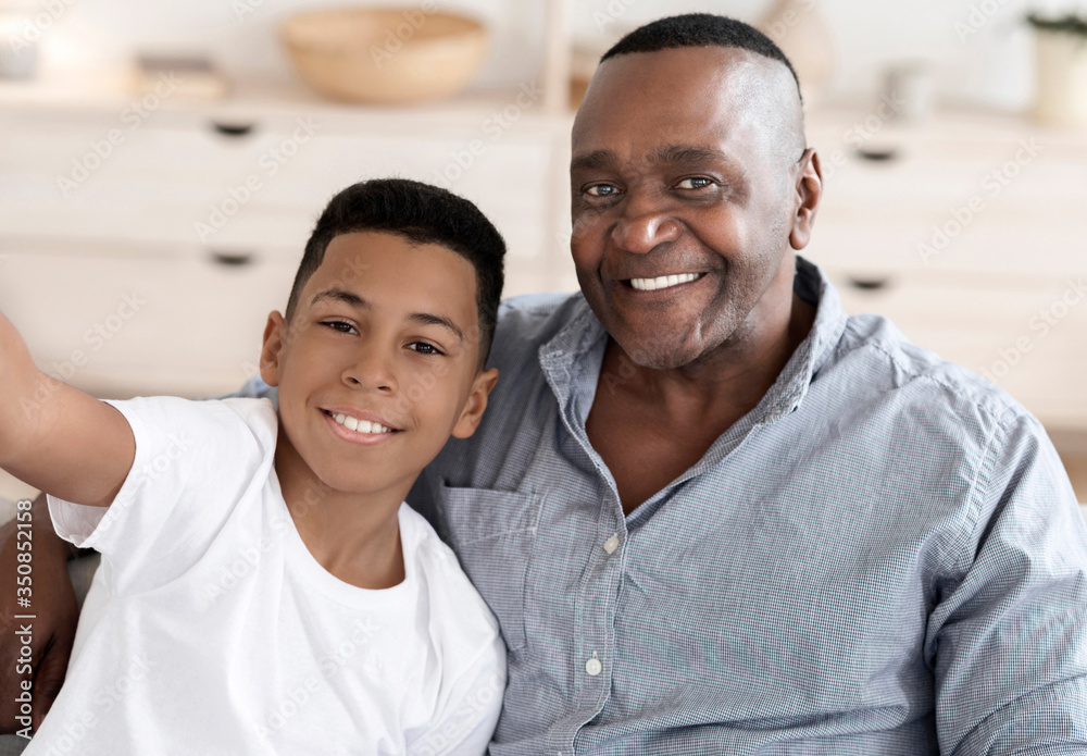 Cheerful black preteen boy taking selfie with his smiling granddad at home Cheerful black preteen boy taking selfie with his smiling granddad at home