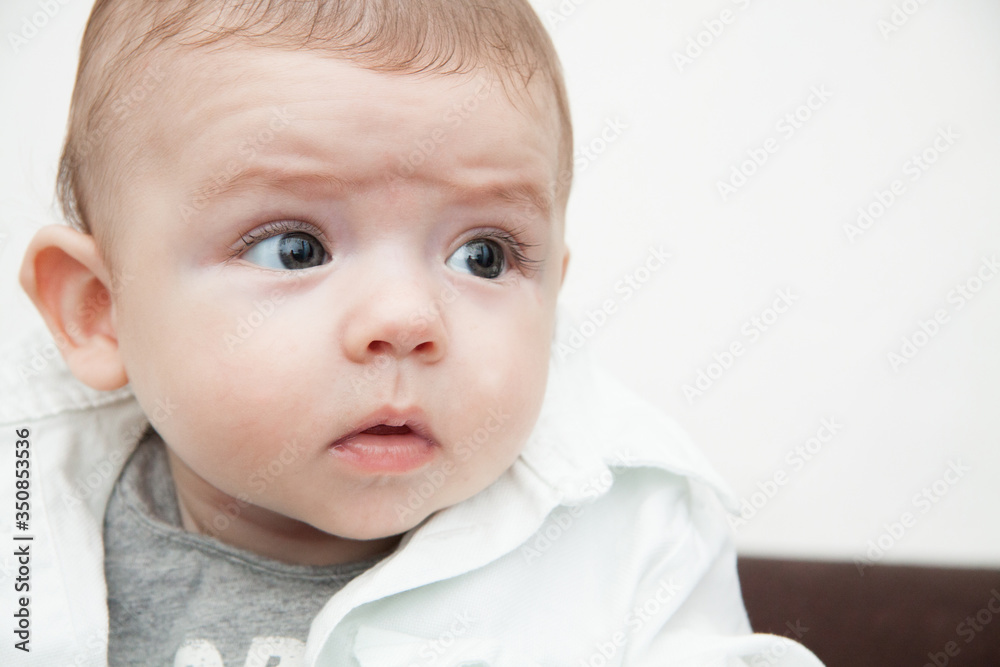 the baby is sitting in a white shirt and looks at the frame. Curious and intelligent children's eyes. Large portrait of a boy. Maternal care. Parenting and motherhood