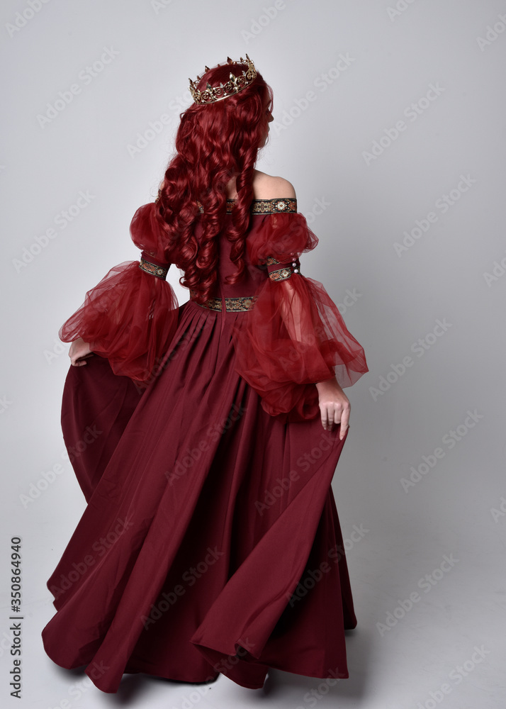 Portrait of a beautiful woman with red hair wearing  a  flowing Burgundy fantasy gown and golden crown.  full length standing pose, isolated against a studio background
