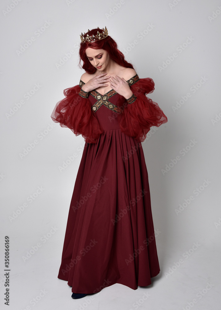 Portrait of a beautiful woman with red hair wearing  a  flowing Burgundy fantasy gown and golden crown.  full length standing pose, isolated against a studio background
