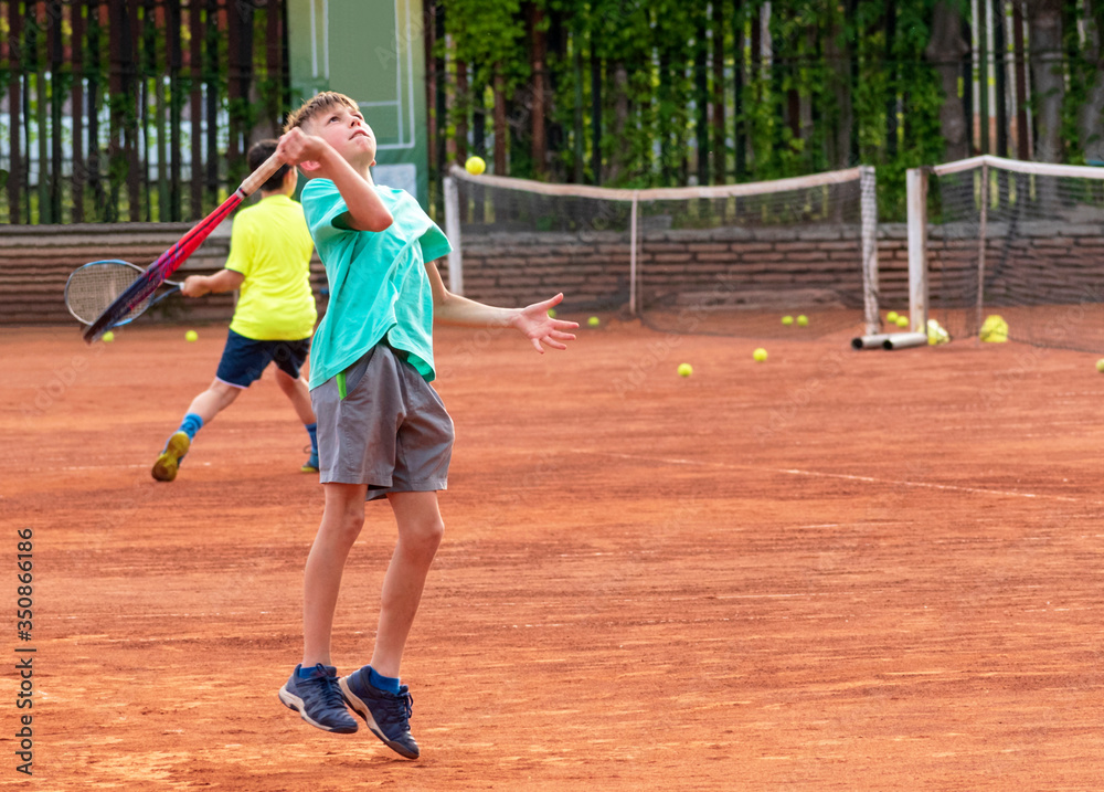 A child tennis player in a jump performs a service at a training session at club. Kids tennis. Boy on the tennis court. Selective focus. Copy space A child tennis player in a jump performs a service at a training session at club. Kids tennis. Boy on the tennis court. Selective focus. Copy space