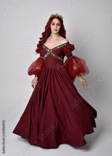 Portrait of a beautiful woman with red hair wearing  a  flowing Burgundy fantasy gown and golden crown.  full length standing pose  isolated against a studio background 