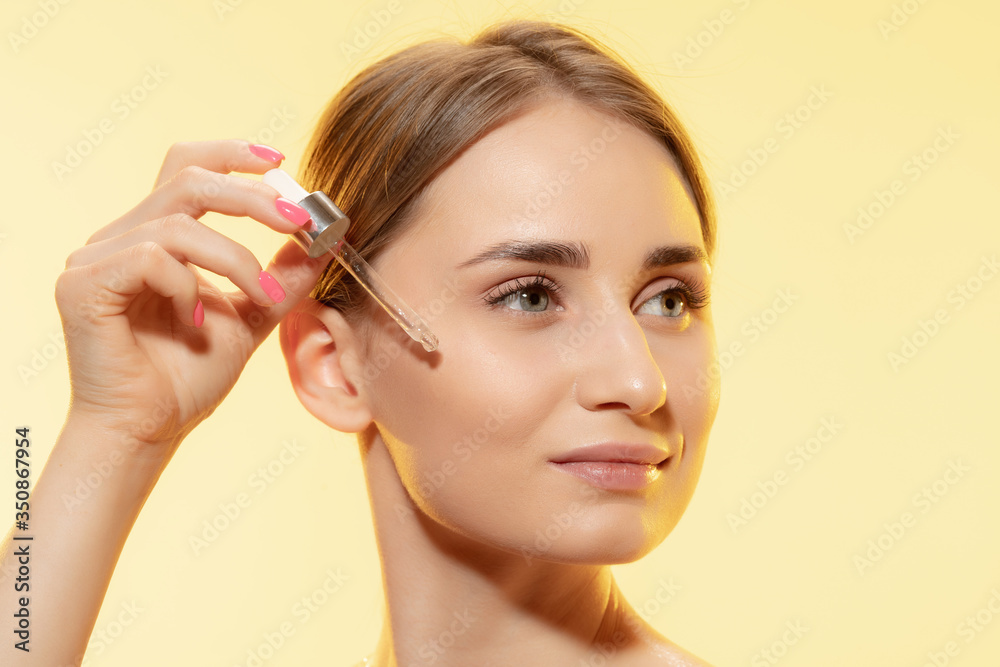 Close up of beautiful young woman with pouring serum, oil on yellow background. Concept of cosmetics, makeup, natural and eco treatment, skin care. Shiny and healthy skin, fashion, healthcare.