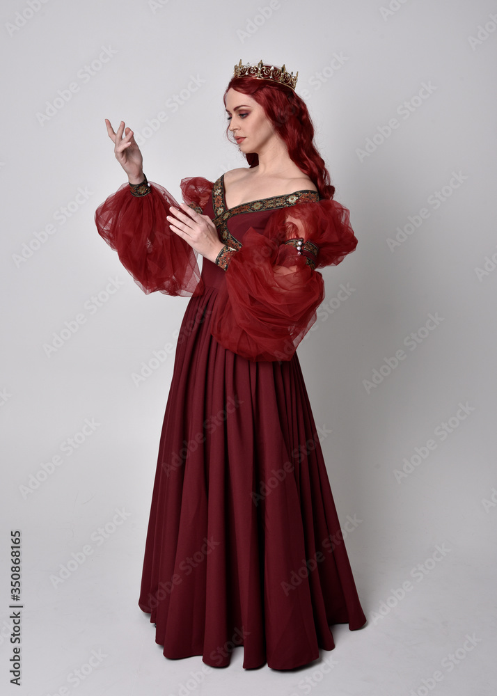 Portrait of a beautiful woman with red hair wearing  a  flowing Burgundy fantasy gown and golden crown.  full length standing pose, isolated against a studio background
