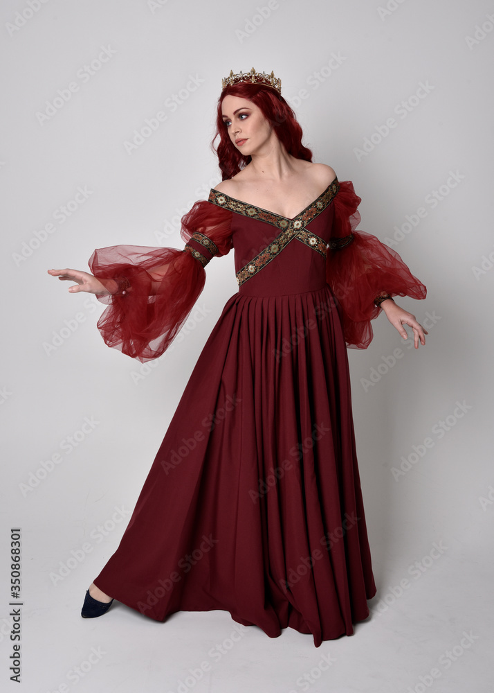 Portrait of a beautiful woman with red hair wearing  a  flowing Burgundy fantasy gown and golden crown.  full length standing pose, isolated against a studio background
