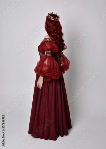 Portrait of a beautiful woman with red hair wearing  a  flowing Burgundy fantasy gown and golden crown.  full length standing pose, isolated against a studio background  © faestock