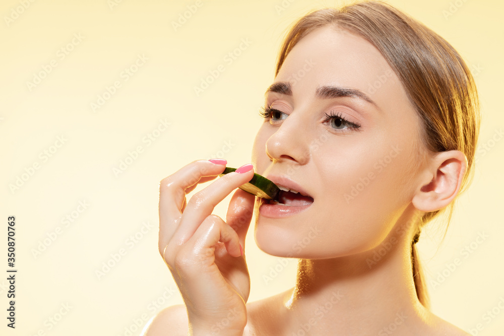 Close up of beautiful young woman with cucumber slice on yellow background. Concept of cosmetics, makeup, natural and eco treatment, skin care. Shiny and healthy skin, fashion, healthcare.