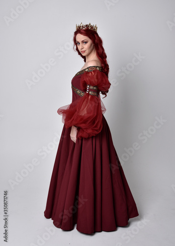 Portrait of a beautiful woman with red hair wearing a flowing Burgundy fantasy gown and golden crown. full length standing pose, isolated against a studio background 