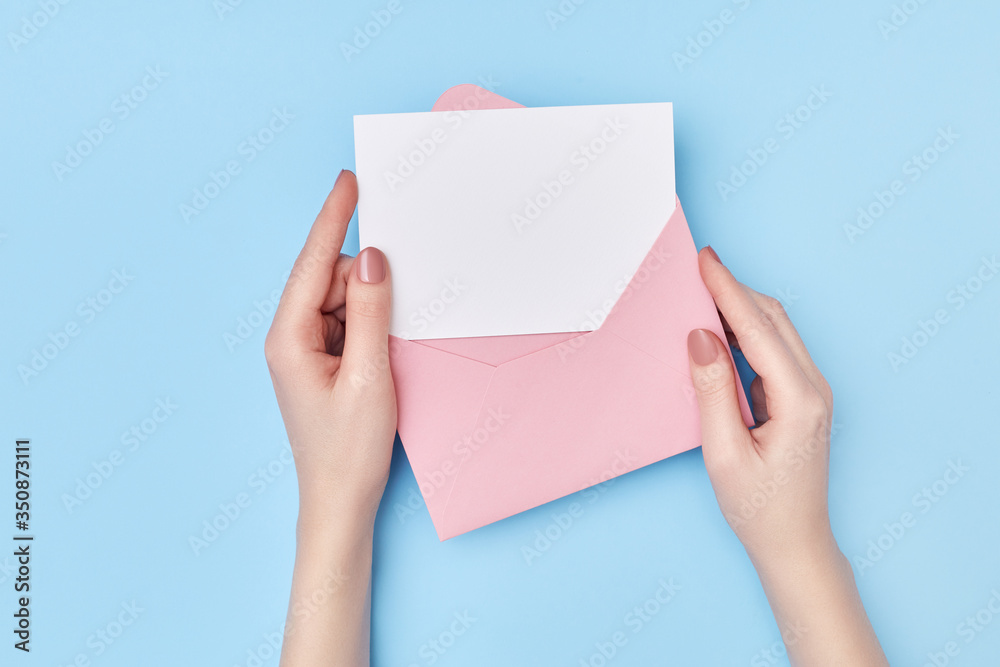 Obraz premium Pink envelope with blank white postcard in woman's hands on the blue background. Mail concept