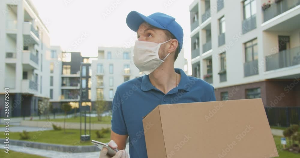 Courier tapping, texting message on smartphone outdoor and looking for address. Delivery man carrying carton box in hands. Courier delivering cardbox parcel during quarantine. Stay home, order online Courier tapping, texting message on smartphone outdoor and looking for address. Delivery man carrying carton box in hands. Courier delivering cardbox parcel during quarantine. Stay home, order online