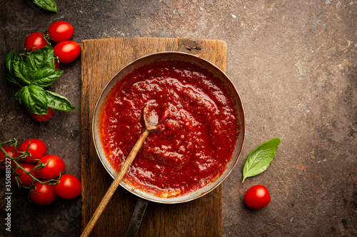 Classic homemade Italian tomato sauce with basil for pasta and pizza in the pan on a wooden chopping board on brown background, top view.