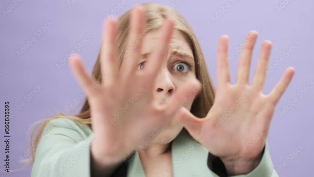 A close-up view of a scared young woman is hiding her face while looking to the camera standing isolated over gray background