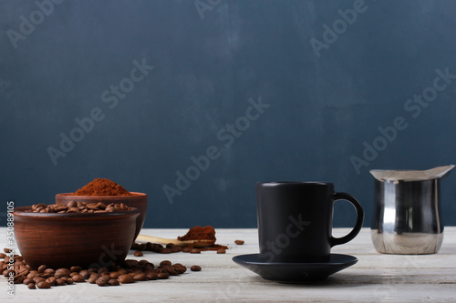 Espresso cup, roasted Arabica beans in clay bowls, coffee powder, metal Turkish pot on while table against dark blue wall. Side view, copy space. Coffee shop, morning, baristas workplace concept