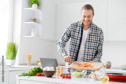 Portrait of positive cheerful gourmet guy enjoy proper nutrition weekend cook fresh salad add sliced tomato wear casual checkered plaid shirt in kitchen house indoors