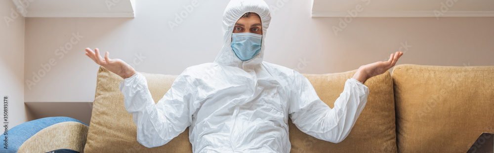 horizontal image of confused man in hazmat suit and protective mask sitting on sofa and showing shrug gesture