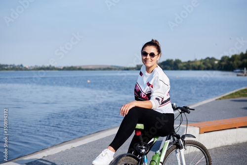 Sep 7, 2019-Ternopil/Ukraine:Young brunette woman, wearing black leggings and white sweatshot, leaning on bicycle. Sports bike ride in city park by lake in summer morning.Healthy active life concept © Natalia