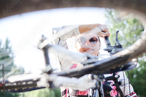 Sep 7, 2019-Ternopil/Ukraine:Young brunette woman, wearing white sweatshot and sunglasses holding bicycle. Sports bike ride in city park by lake in summer morning. Healthy active life concept. photo