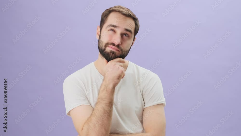 An attractive young man wearing a white t-shirt is dreaming while touching his beard and smiling standing isolated over gray background An attractive young man wearing a white t-shirt is dreaming while touching his beard and smiling standing isolated over gray background