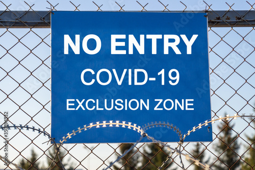 The inscription "NO ENTRY covid-19 exclusion zone" on a blue plate on an iron mesh fence rewound with barbed wire. Closed area. Quarantine and lockdown concept