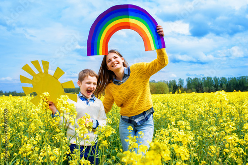 Child holding drawing of colorful rainbow, closeup. Adaptation concept. World Adoption Day September 30th. Yellow field of blooming rapeseed, blue sky and clouds. Freedom childhood and summer photo